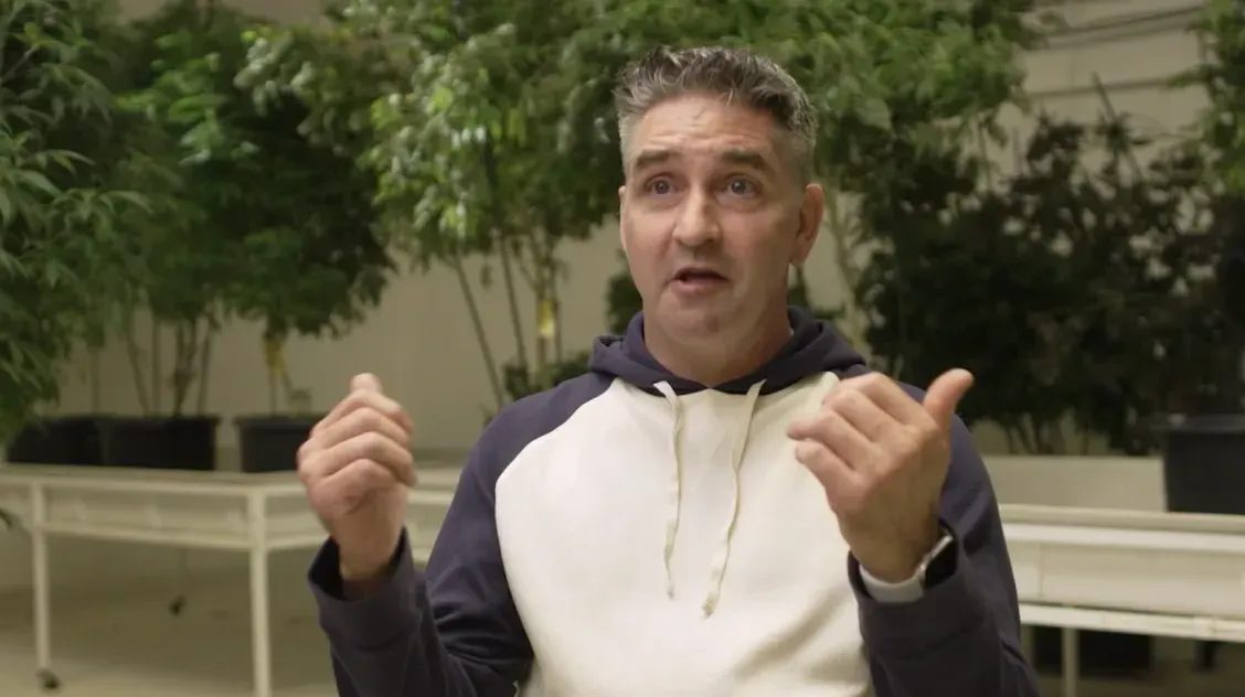 Man speaking with hands raised in front of indoor cannabis plants